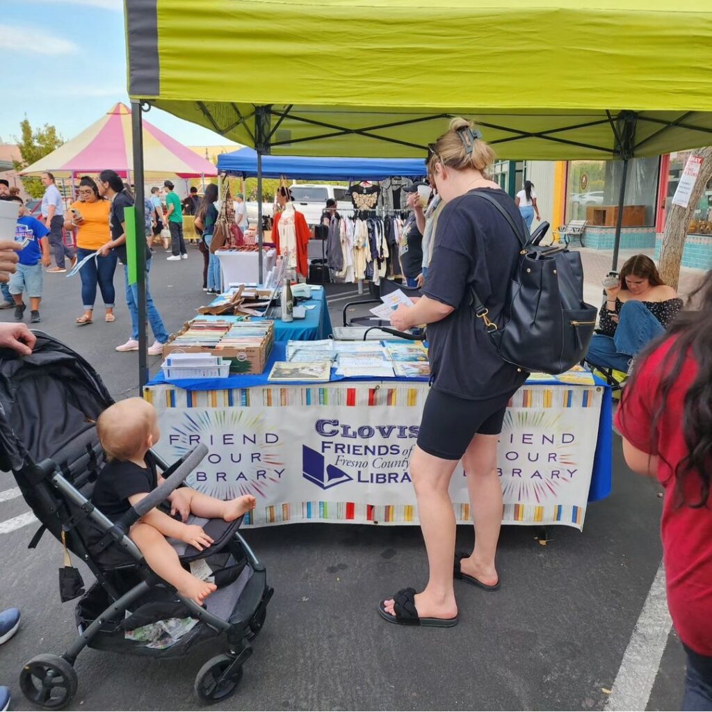 A woman and a baby in a stroller examine the books available at the Clovis Friends of the Library table at the Clovis Farmer's Market.