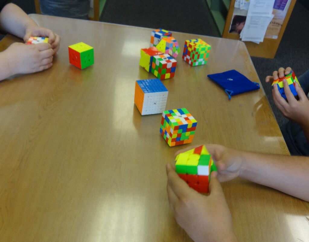 The hands of three kids can be seen playing with brightly-colored cubes during a Cubing Club meeting. There are eleven cubes on the table, all in different colors.