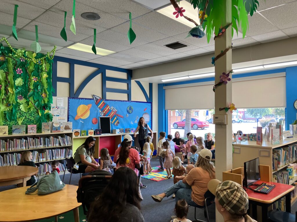 Claire Noland leads a storytime in the Children's Area of the Kingsburg branch. Several parents are seated around the room, some of them with their smaller children, while most of the children engage in an activity. The area is decorated with paper plants and a space-themed background.