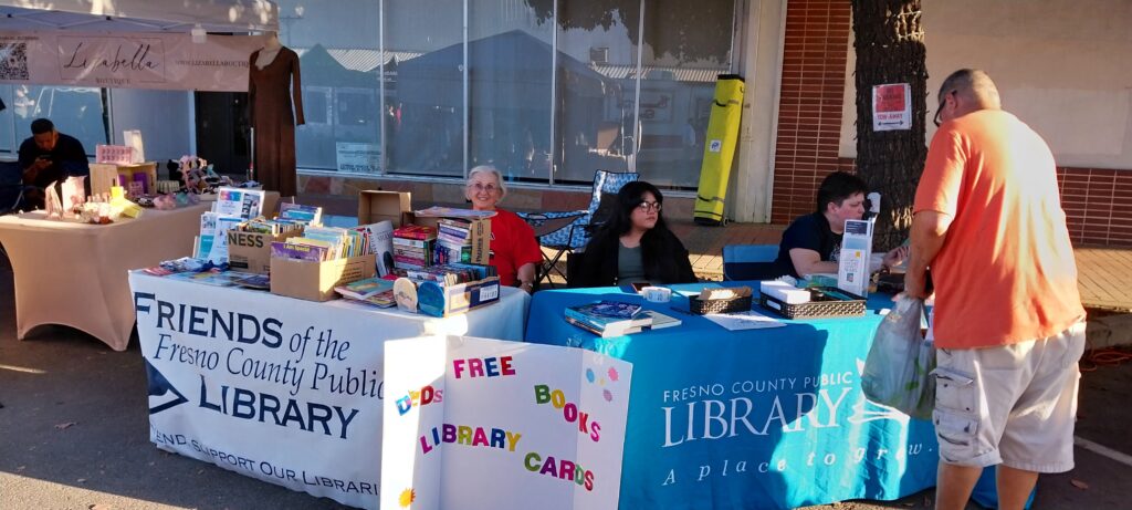 Sanger FOL President Rosa Huerta sits behind a Friends of the Library table, next to a Fresno County Public Library table. Her table is loaded with books for sale, as well as fliers and promotional materials.