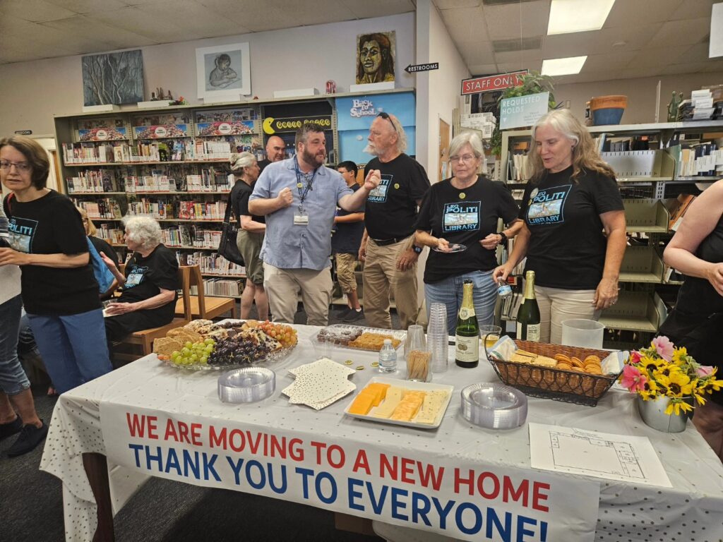 Several members of the Politi Friends wear black Leo Politi Library t-shirts and stand behind a table with a banner that reads: "We are moving to a new home; thank you to everyone." On the table, there are two bottles of sparkling cider, as well as a spread of grapes, cheese, crackers, cookies, and other snacks.