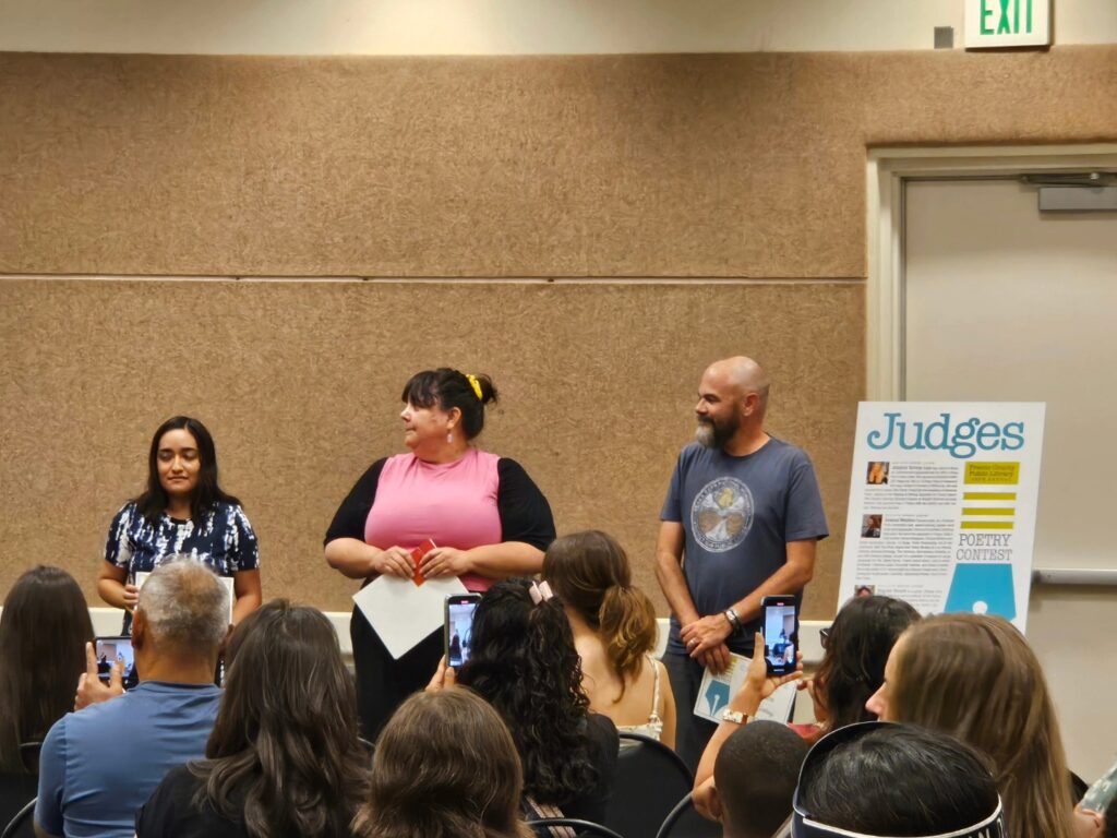 The three adult Poetry Contest winners stand at the front of the Meeting Room at the Woodward Park branch to accept their awards while the audience takes photos.