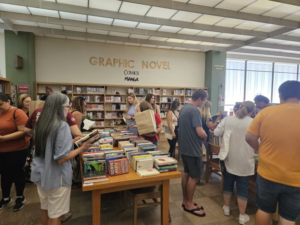 Over a dozen people browse the tables and carts of books for sale in the Graphic Novel section of the Central Library.